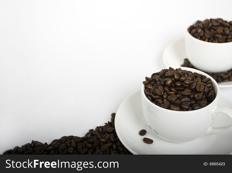 Two coffee cups filled with espresso coffee beans on the right-hand side of a white frame. Beans placed around saucers and a border of beans at the bottom. Two coffee cups filled with espresso coffee beans on the right-hand side of a white frame. Beans placed around saucers and a border of beans at the bottom.