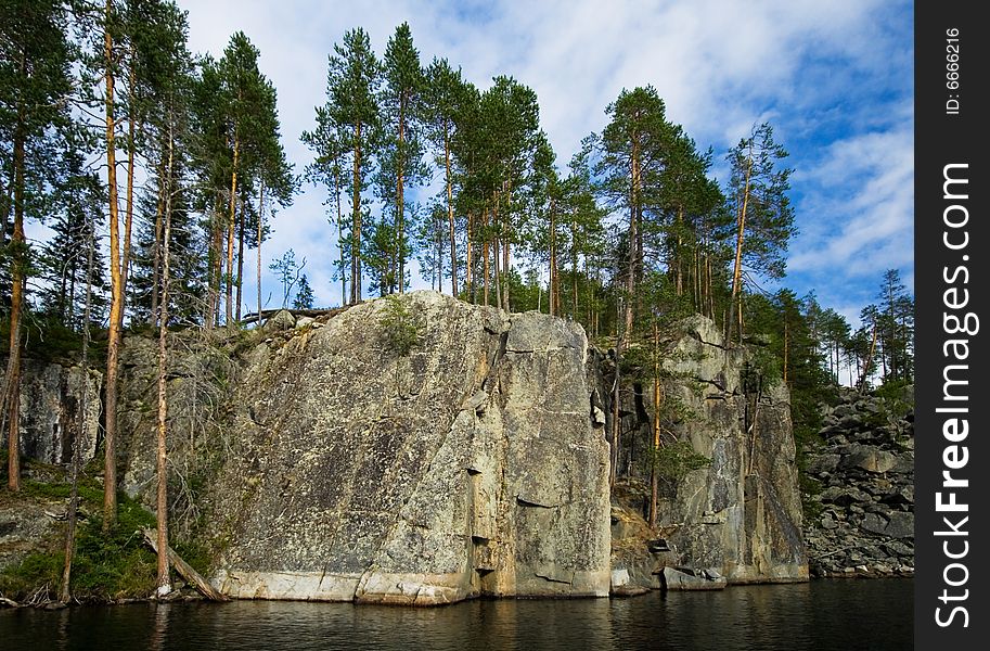A wild sheer cliff rises from the black water of Karelia Pizanets lake with pine tress on it. A wild sheer cliff rises from the black water of Karelia Pizanets lake with pine tress on it