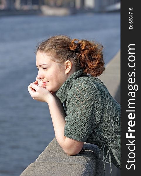 Old-fashioned Young curly girl in a vintage gray knitted dress stands on the promenade and looks at the river. She smiles slightly. Old-fashioned Young curly girl in a vintage gray knitted dress stands on the promenade and looks at the river. She smiles slightly.