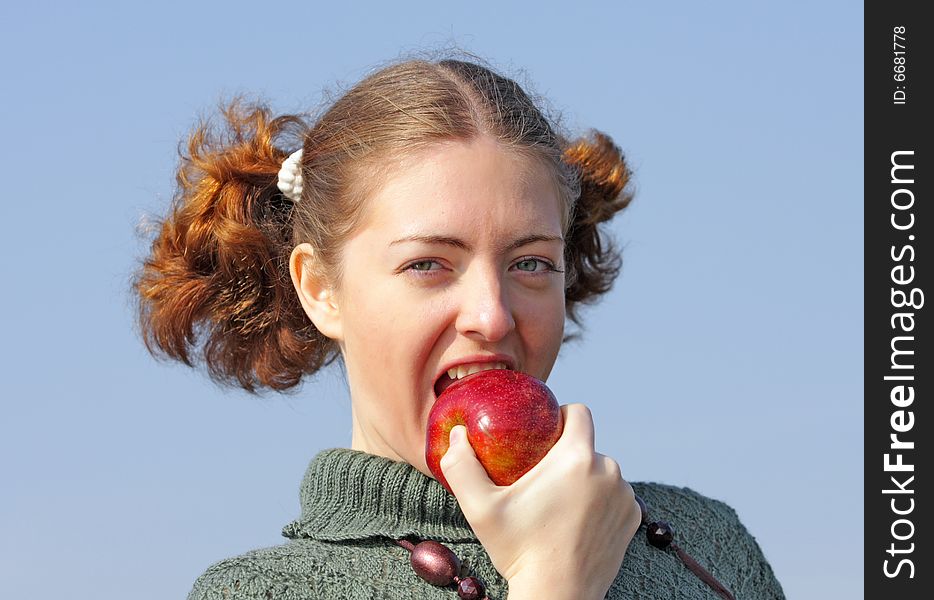 Old-fashioned Young curly girl in a vintage gray knitted dress bites a big red apple. The woman is a vegan. Blue sky background. Pretty girl is eating apple