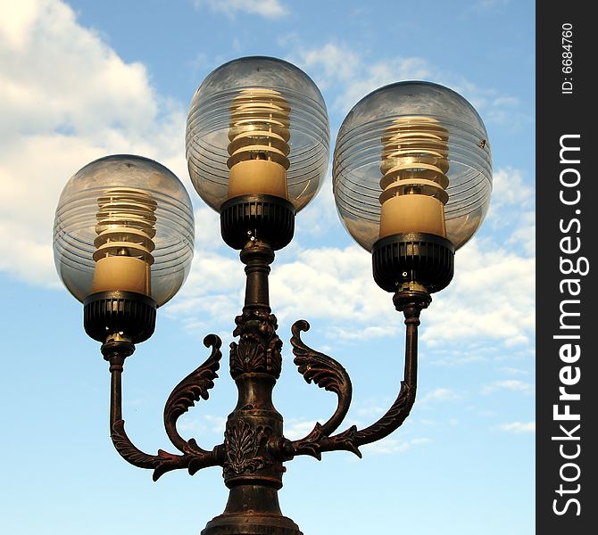 Three ornate street lamps on a lamp post against a blue sky in Romania