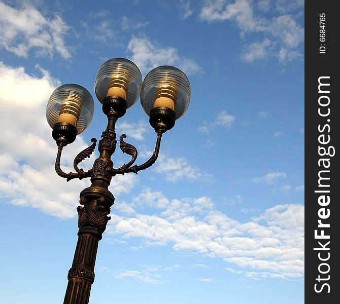 Three ornate street lamps on a lamp post against a blue sky in Romania