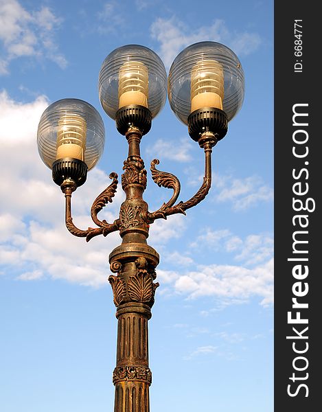 Three ornate street lamps on a lamp post against a blue sky in Romania