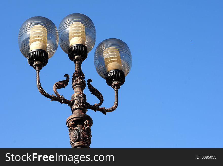 Three ornate street lamps on a lamp post against a blue sky in Romania