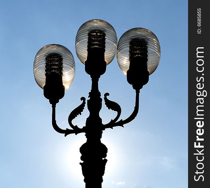 Three ornate street lamps on a lamp post against a blue sky in Romania