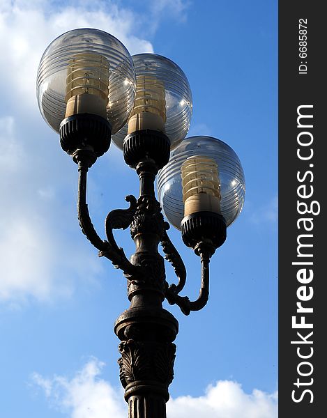 Three ornate street lamps on a lamp post against a blue sky in Romania