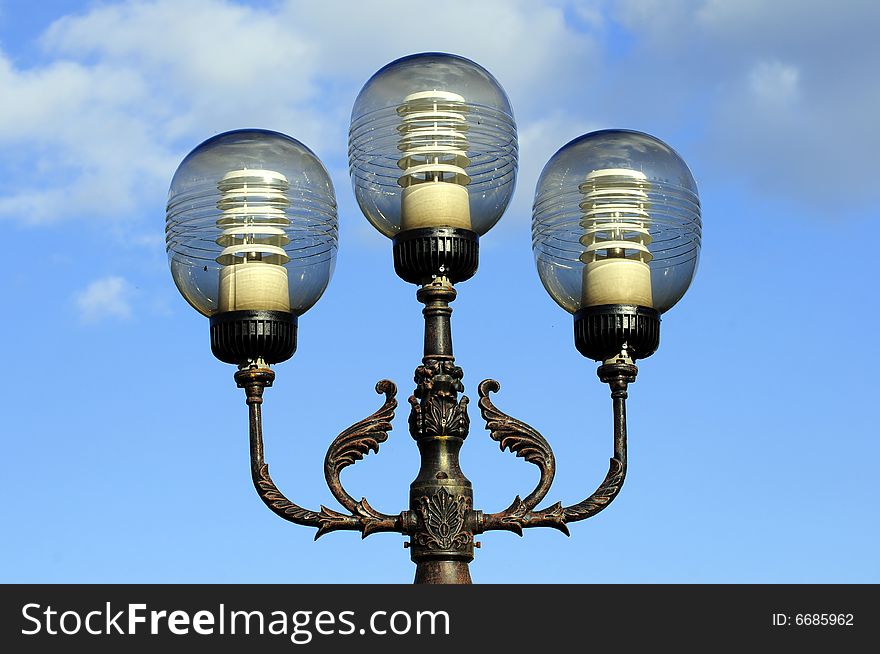 Three ornate street lamps on a lamp post against a blue sky in Romania