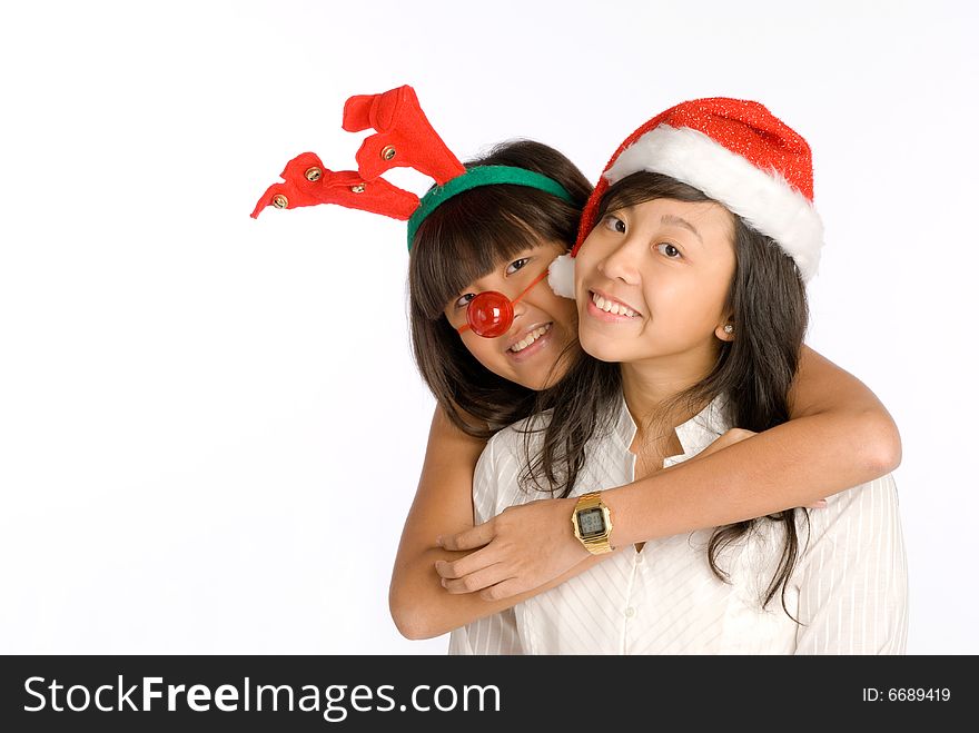 Tall lanky teenager Asian girl in antlers head wear and red nose hugging shorter girl in Santa Claus hat from behind at a Christmas party. Tall lanky teenager Asian girl in antlers head wear and red nose hugging shorter girl in Santa Claus hat from behind at a Christmas party.