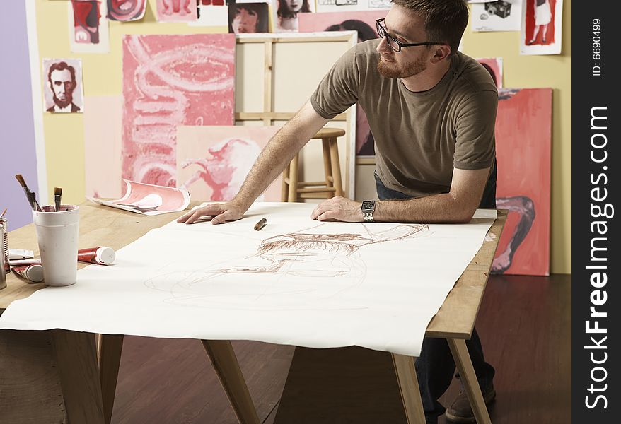 Artist leans over a artwork and is drawing with a brown crayon.  He looks to the left of camera. Artist leans over a artwork and is drawing with a brown crayon.  He looks to the left of camera.