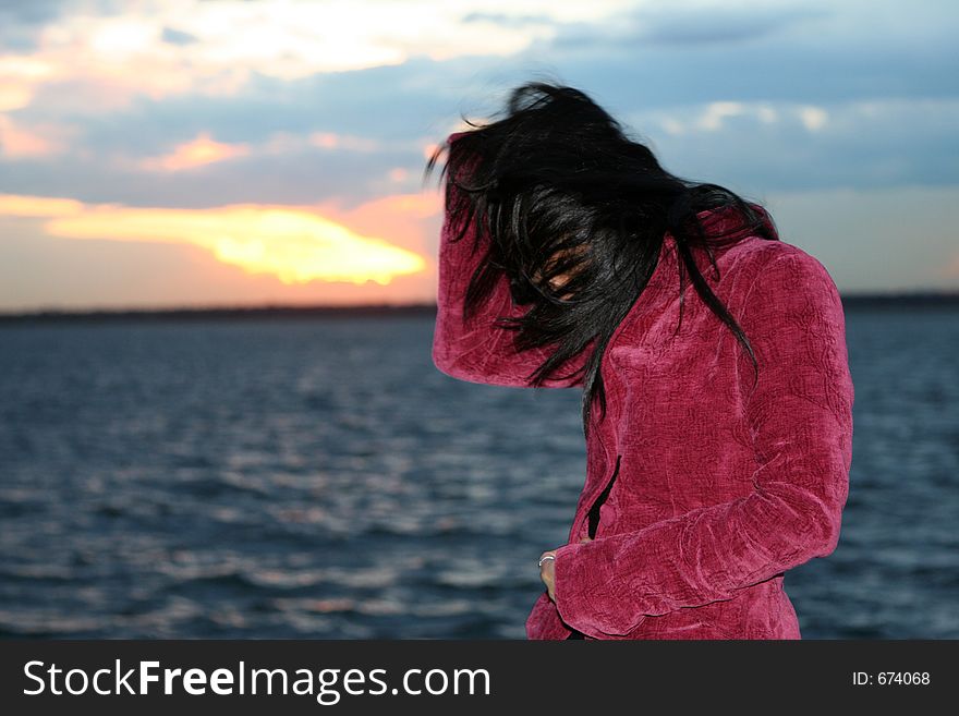 A girl in a jacket by the water at the sunset. Wind is blowing hair in her face. A girl in a jacket by the water at the sunset. Wind is blowing hair in her face.