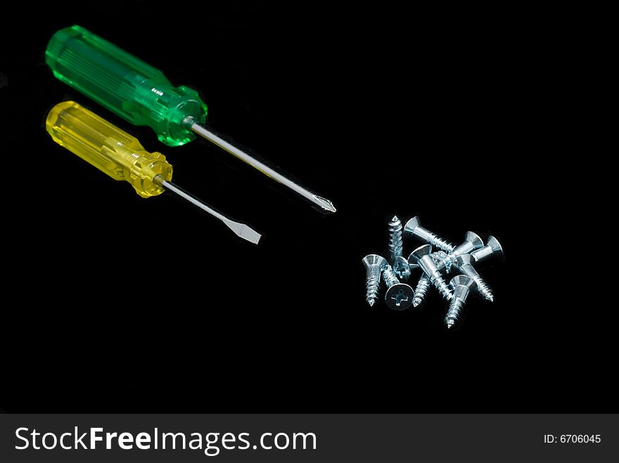 A yellow and a green screwdriver and a pile of screws on a black background. A yellow and a green screwdriver and a pile of screws on a black background