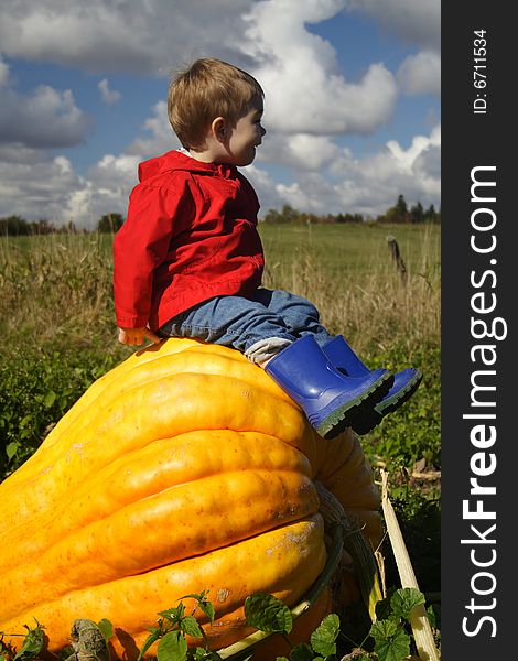 A young boy wearing a red coat and blue rubber boots sitting proudly on top of a big orange pumpkin. A young boy wearing a red coat and blue rubber boots sitting proudly on top of a big orange pumpkin.