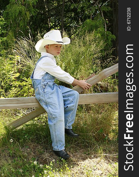 Young boy sitting on a broken fence wearing overall and a cowboy hat. Young boy sitting on a broken fence wearing overall and a cowboy hat