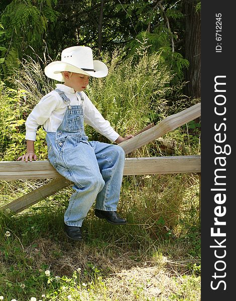 Young boy sitting on a broken fence wearing overall and a cowboy hat. Young boy sitting on a broken fence wearing overall and a cowboy hat