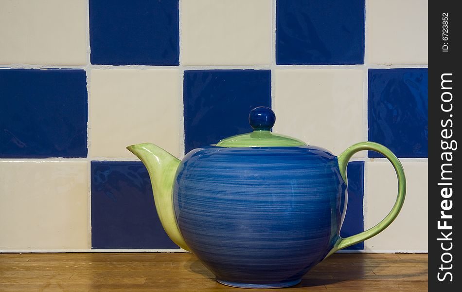 A cheery blue and green teapot stands on a wood counter in front of blue and white tiles. A cheery blue and green teapot stands on a wood counter in front of blue and white tiles.