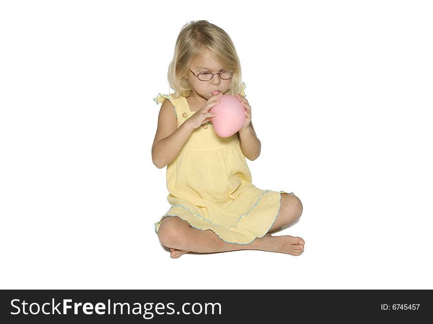 A young girl in a yellow dress sitting down and blowing up a pink balloon. Isolated on a white background. A young girl in a yellow dress sitting down and blowing up a pink balloon. Isolated on a white background