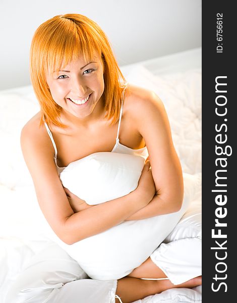 Young red-haired woman dressed pyjamas sitting on bed and hugging pillow. She's smiling and looking at camera. Front view. Young red-haired woman dressed pyjamas sitting on bed and hugging pillow. She's smiling and looking at camera. Front view.