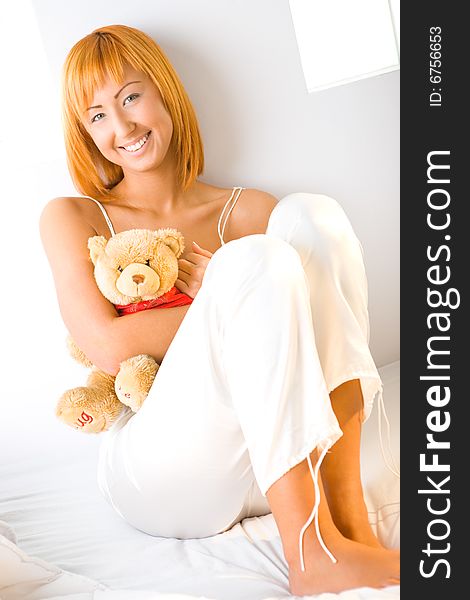 Young red-haired woman dressed pyjamas sitting on bed and hugging teddy bear. She's leaning against a wall. She's smiling and looking at camera. Young red-haired woman dressed pyjamas sitting on bed and hugging teddy bear. She's leaning against a wall. She's smiling and looking at camera.