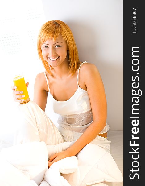 Young red-haired woman dressed pyjamas sitting on bed with glass of juice. She's leaning against a wall. She's smiling and looking at camera. Young red-haired woman dressed pyjamas sitting on bed with glass of juice. She's leaning against a wall. She's smiling and looking at camera.