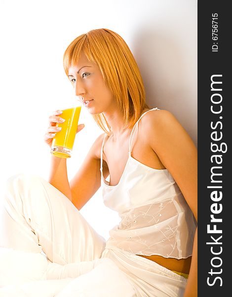 Young red-haired woman dressed pyjamas sitting on bed with glass of juice. She's leaning against a wall. Young red-haired woman dressed pyjamas sitting on bed with glass of juice. She's leaning against a wall.