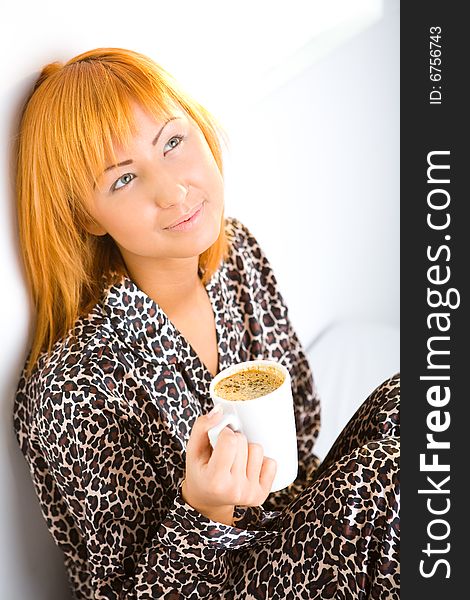 Young red-haired woman dressed pyjamas sitting on bed with cup of coffee. She's leaning against a wall. Young red-haired woman dressed pyjamas sitting on bed with cup of coffee. She's leaning against a wall.