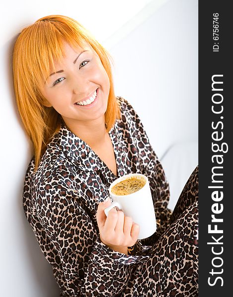 Young red-haired woman dressed pyjamas sitting on bed with cup of coffee. She's leaning against a wall. She's smiling and looking at camera. Young red-haired woman dressed pyjamas sitting on bed with cup of coffee. She's leaning against a wall. She's smiling and looking at camera.