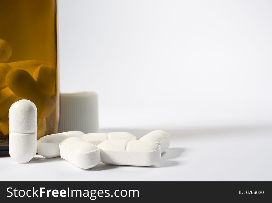 A group of pills on the lower left-hand side of a white frame with one standing up next to a brown bottle. A group of pills on the lower left-hand side of a white frame with one standing up next to a brown bottle