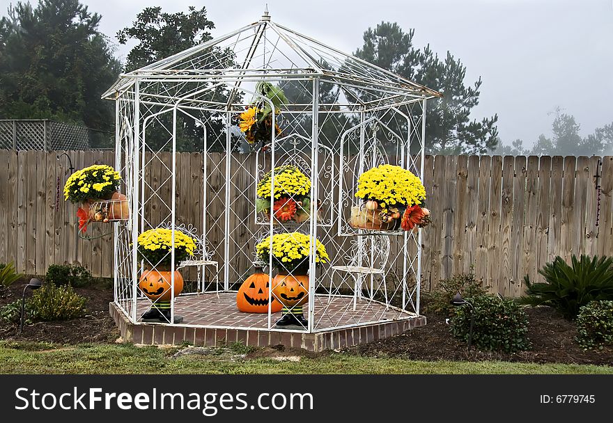 A white iron gazebo decorated for fall with mums, pumpkins and jack-o-laterns overcast by a hazy sky. A white iron gazebo decorated for fall with mums, pumpkins and jack-o-laterns overcast by a hazy sky.