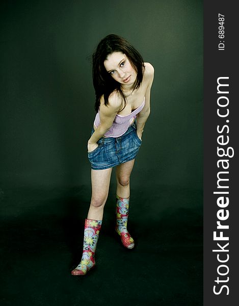 Young pretty brunette poses in the studio, dressed casual, with a light lilac top, denim skirt and colorful, funny plastic boots, trying various expressions, over a black background. Young pretty brunette poses in the studio, dressed casual, with a light lilac top, denim skirt and colorful, funny plastic boots, trying various expressions, over a black background