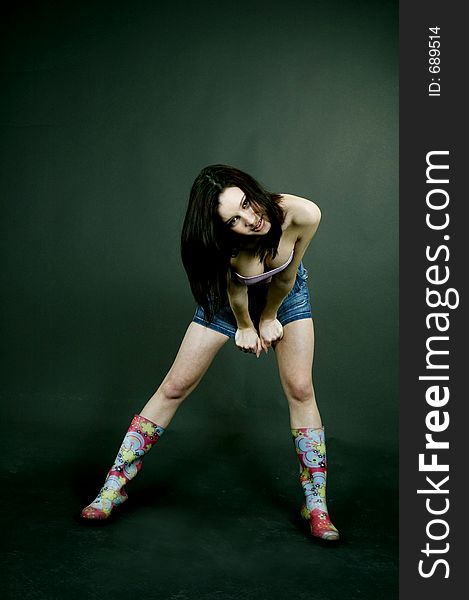Young pretty brunette poses in the studio, dressed casual, with a light lilac top, denim skirt and colorful, funny plastic boots, trying various expressions, over a black background. Young pretty brunette poses in the studio, dressed casual, with a light lilac top, denim skirt and colorful, funny plastic boots, trying various expressions, over a black background