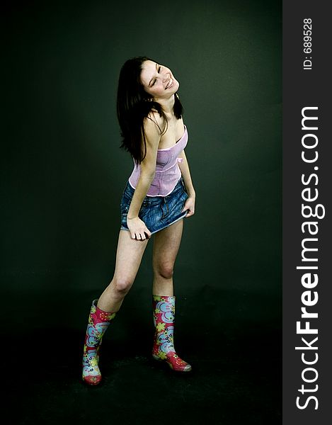 Young pretty brunette poses in the studio, dressed casual, with a light lilac top, denim skirt and colorful, funny plastic boots, trying various expressions, over a black background. Young pretty brunette poses in the studio, dressed casual, with a light lilac top, denim skirt and colorful, funny plastic boots, trying various expressions, over a black background