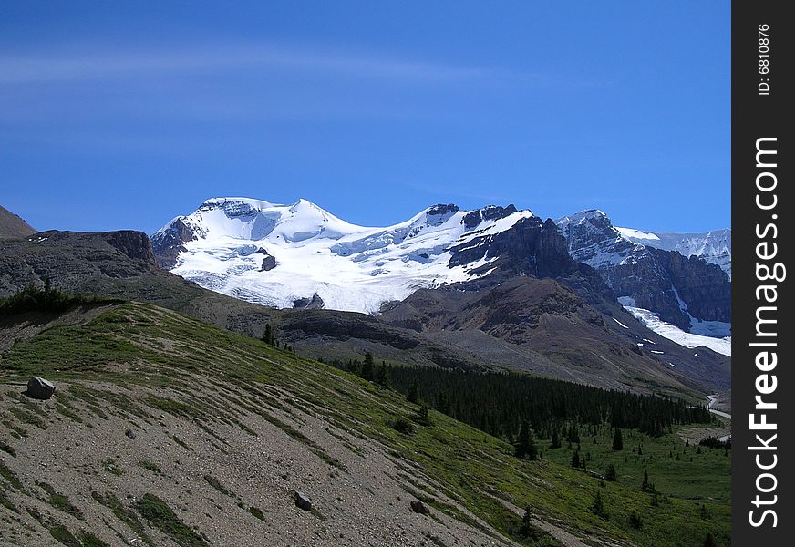 A snow covered Canadian mountain framed by meadows and a gravelly slope.