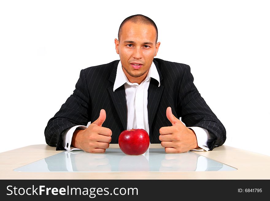 Young businessman sitting in front of a red apple thinking with his thumbs up. Isolate over white. The table mirrors the image creating a nice effect!. Young businessman sitting in front of a red apple thinking with his thumbs up. Isolate over white. The table mirrors the image creating a nice effect!