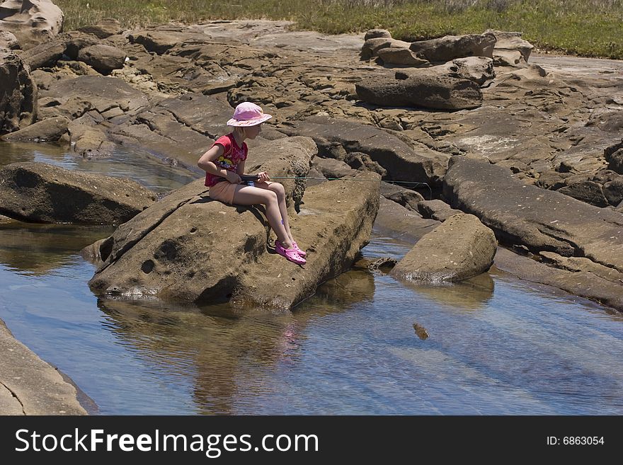 Little girl fishing in a rock pool with a fishing rod in bright sunshine dressed in bright pink and red