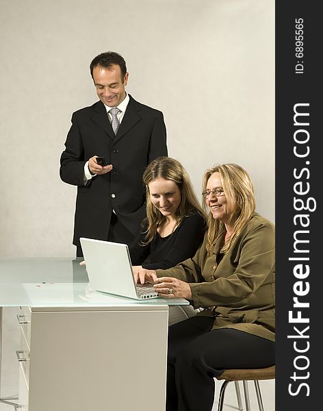 Two women seated at a desk looking at a computer and smiling while a man stands nearby looking at his cell phone. Vertically framed photo. Two women seated at a desk looking at a computer and smiling while a man stands nearby looking at his cell phone. Vertically framed photo.