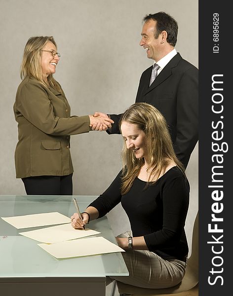 Three people are in a room together. The youngest member is smiling and studying paper on the table in front of her. The other two are shaking hands and smiling at each other. Vertically framed shot. Three people are in a room together. The youngest member is smiling and studying paper on the table in front of her. The other two are shaking hands and smiling at each other. Vertically framed shot.