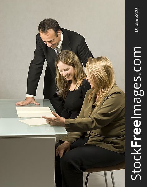 Three people are in a business meeting. They are smiling and looking at some pieces of paper on the table. The women are sitting and the man is standing. Vertically framed shot. Three people are in a business meeting. They are smiling and looking at some pieces of paper on the table. The women are sitting and the man is standing. Vertically framed shot.