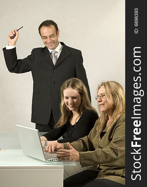 Two women at a desk in front of a laptop shaking hands and smiling, while a man stands watching and smiling. Vertically framed photo. Two women at a desk in front of a laptop shaking hands and smiling, while a man stands watching and smiling. Vertically framed photo.