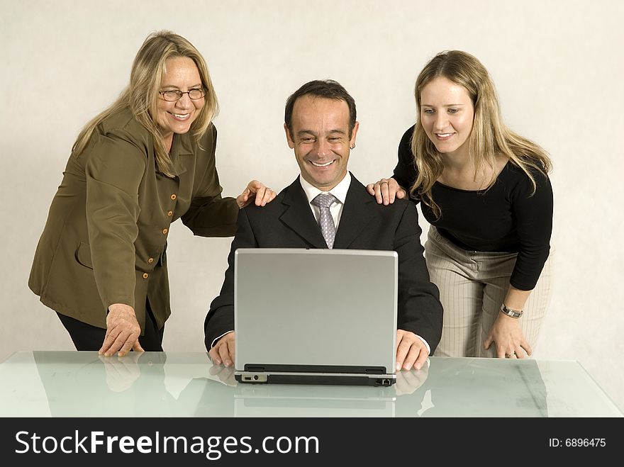 Two women standing over a man seated at adesk with their hands upon his shoulders as they all smile. Horizontally framed photo. Two women standing over a man seated at adesk with their hands upon his shoulders as they all smile. Horizontally framed photo.