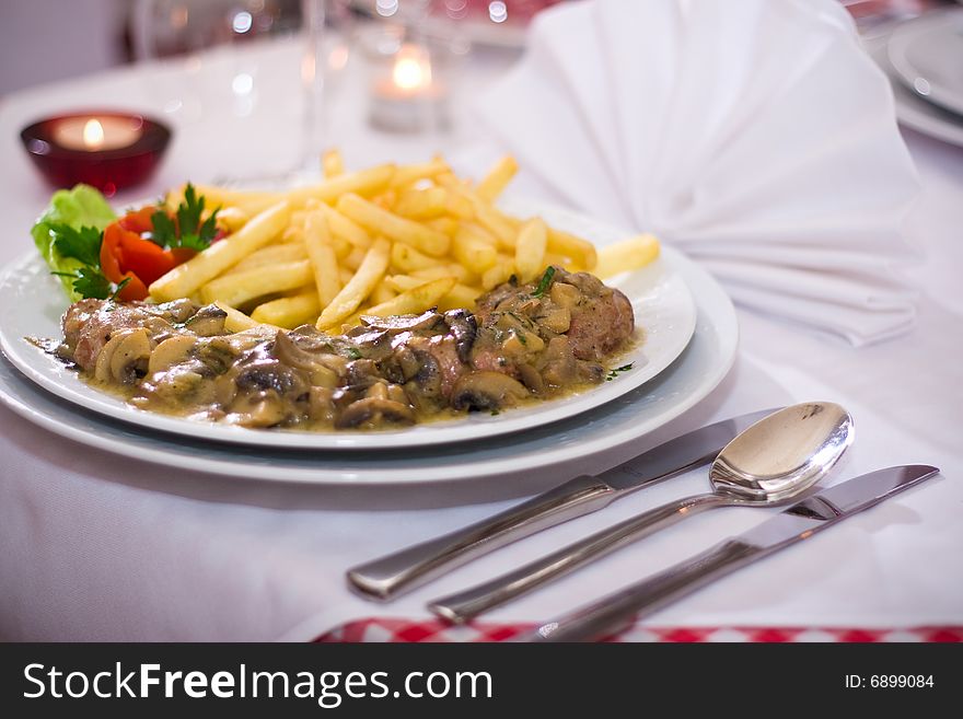 French fries with mushrooms and pork meat in the background are candles, cutlery and napkins. French fries with mushrooms and pork meat in the background are candles, cutlery and napkins