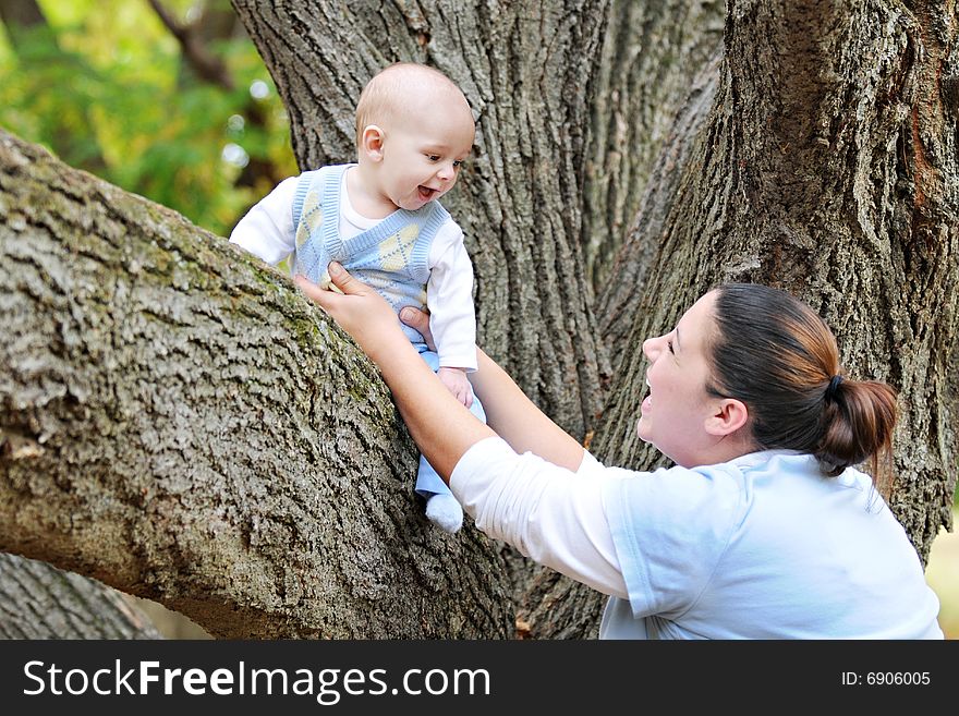 A baby sitting on a big tree limb, laughing at his mother who's securing him. A baby sitting on a big tree limb, laughing at his mother who's securing him.