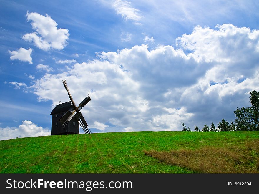 Wooden windmill against the summer blue sky with white clouds on a green hill with a birch