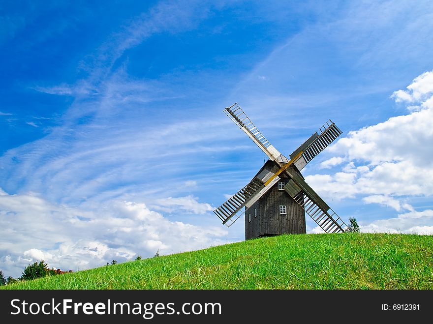 Wooden windmill against the summer blue sky with white clouds on a green hill with a birch