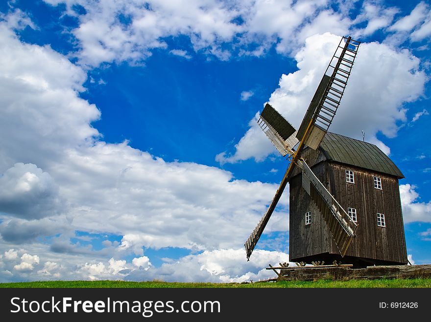 Wooden windmill against the summer blue sky with white clouds on a green hill with a birch