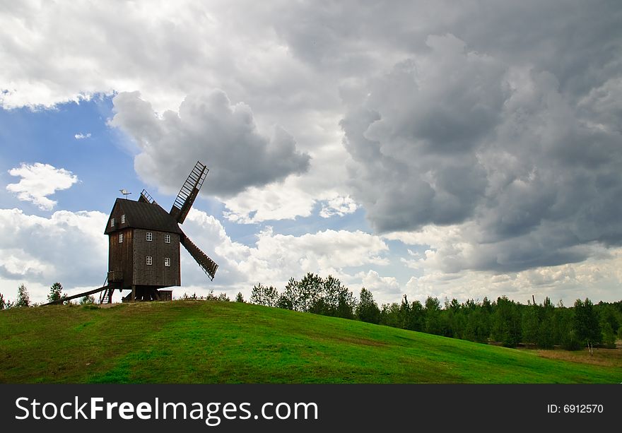 Wooden windmill against the summer blue sky with white clouds on a green hill with a birch