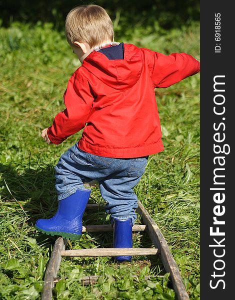 A toddler wearing a red jacket and blue rubber boots walking over a ladder in the apple orchard. A toddler wearing a red jacket and blue rubber boots walking over a ladder in the apple orchard.