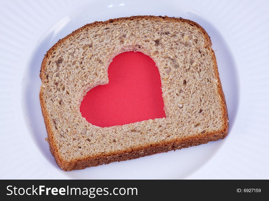 Red heart pattern cut in a slice of whole grain bread, on a white plate. Red heart pattern cut in a slice of whole grain bread, on a white plate.