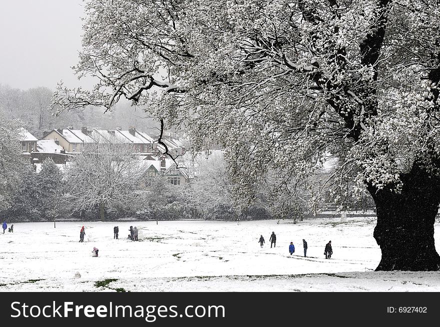Winter’s scene with snow falling people and children playing and framed by a prominent tree with snowy covered branches. Winter’s scene with snow falling people and children playing and framed by a prominent tree with snowy covered branches.