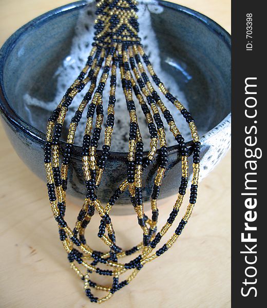 A black and gold beaded necklace presented over a ceramic bowl. This is an example of the craft of beading. The focus is on the beads close to the rim of the bowl. A black and gold beaded necklace presented over a ceramic bowl. This is an example of the craft of beading. The focus is on the beads close to the rim of the bowl.