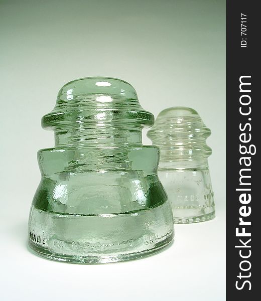 Two old glass insulators, one green and one clear, with light reflecting off them. Focus is on the front insulator. Writing, Made in the USA and numbers. Two old glass insulators, one green and one clear, with light reflecting off them. Focus is on the front insulator. Writing, Made in the USA and numbers.